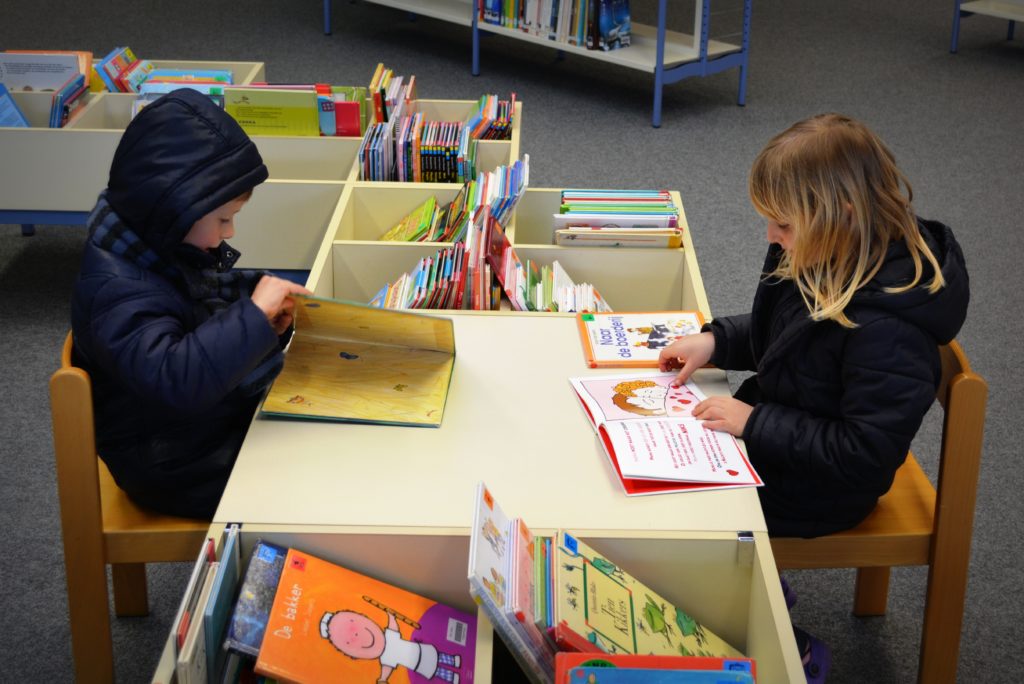 Two young children sit at a library table reading foreign language books