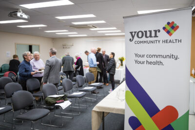 Your Community Health banner in front of room of people meeting for the AGM 2019