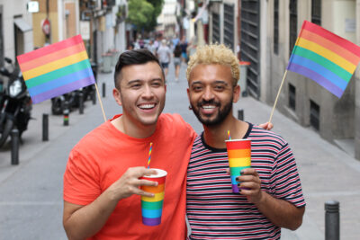 Portrait of two men, arm around each other holding rainbow flags and rainbow cups on a Melbourne street