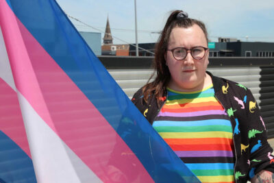 Head and shoulders picture of Elle, outside in a rainbow stripe top, standing in from of a trans flag. redit: ABC News, Darryl Torpy
