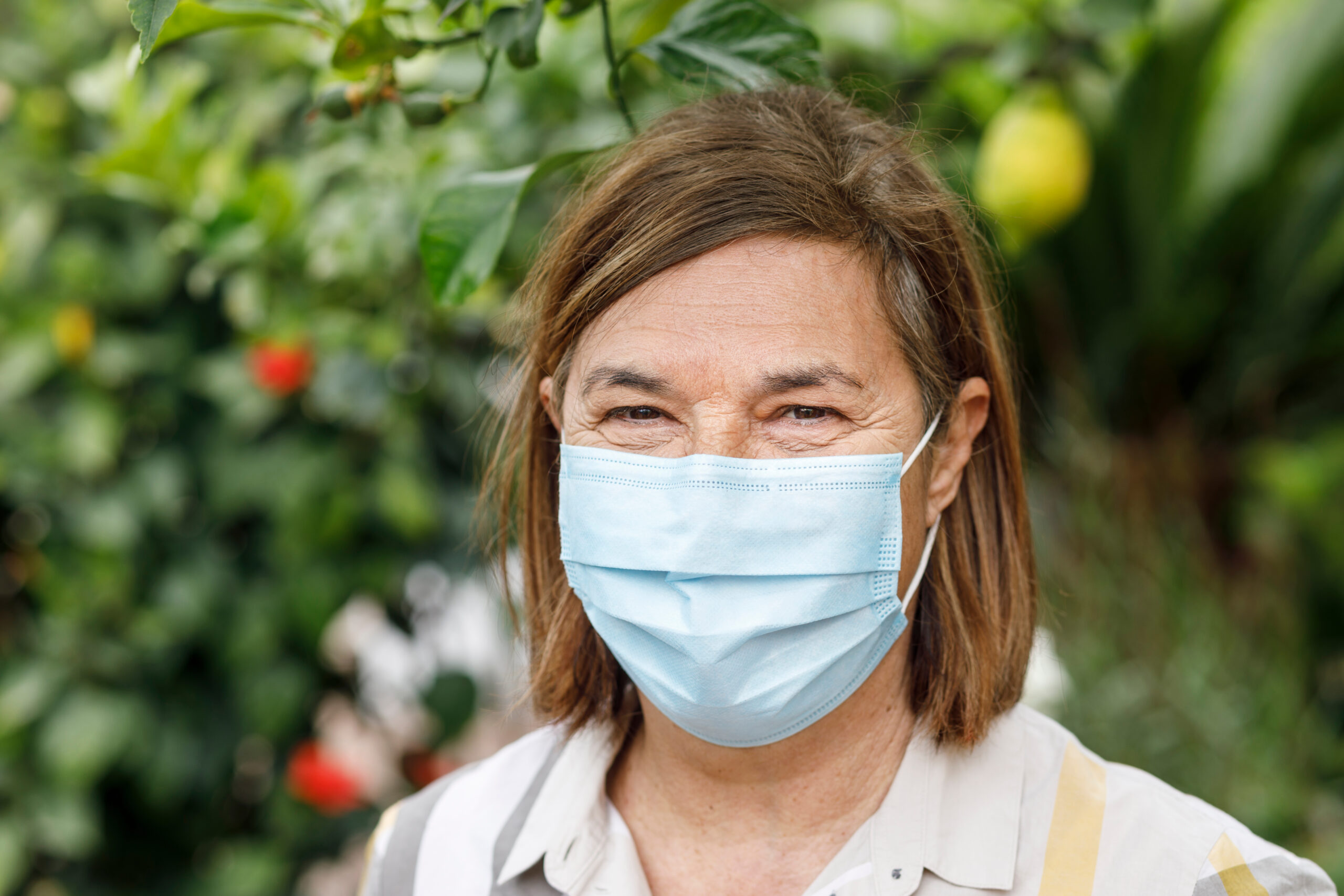 Close up of woman brown bobbed hair wearing a face mask. Blurred green garden background.