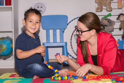 A child sits on the floor holding beads on a string for counting. A woman with glasses and a red top lies on the floor next to him and is handing him a bead.