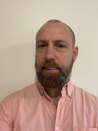 Head shot of Kent, a man with a shaved head and beard in a pink shirt.