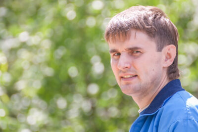 White man in a blue top stands side on and looks at the camera. He is outside and the green background is blurred.