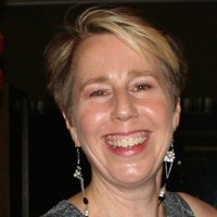 A close up photo of a smiling woman with short blonde hair and long earrings on a black background.