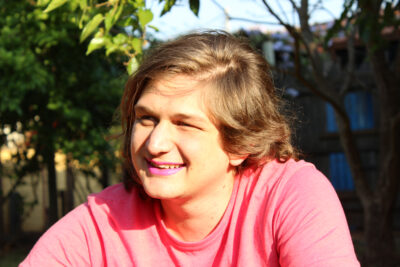 A young gender diverse person, in a pink top and pink lipstick,sitting outside smiling, looking away from the camera.