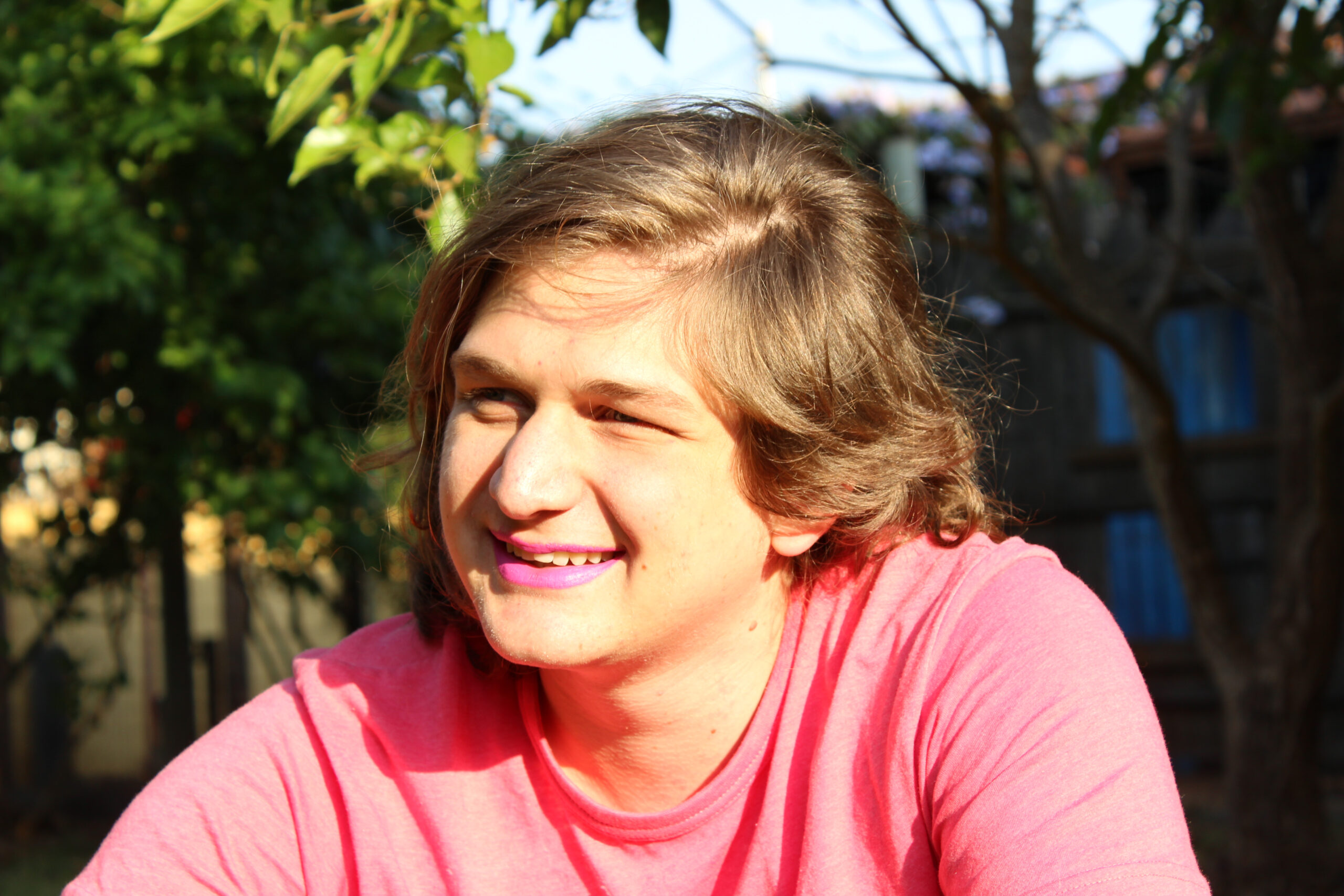 A young trans person, in a pink top and pink lipstick,sitting outside smiling, looking away from the camera.