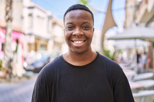 A young, black, trans man smiles at the camera. He is standing on a street wearing a black tshirt, short hair and a nose piercing.